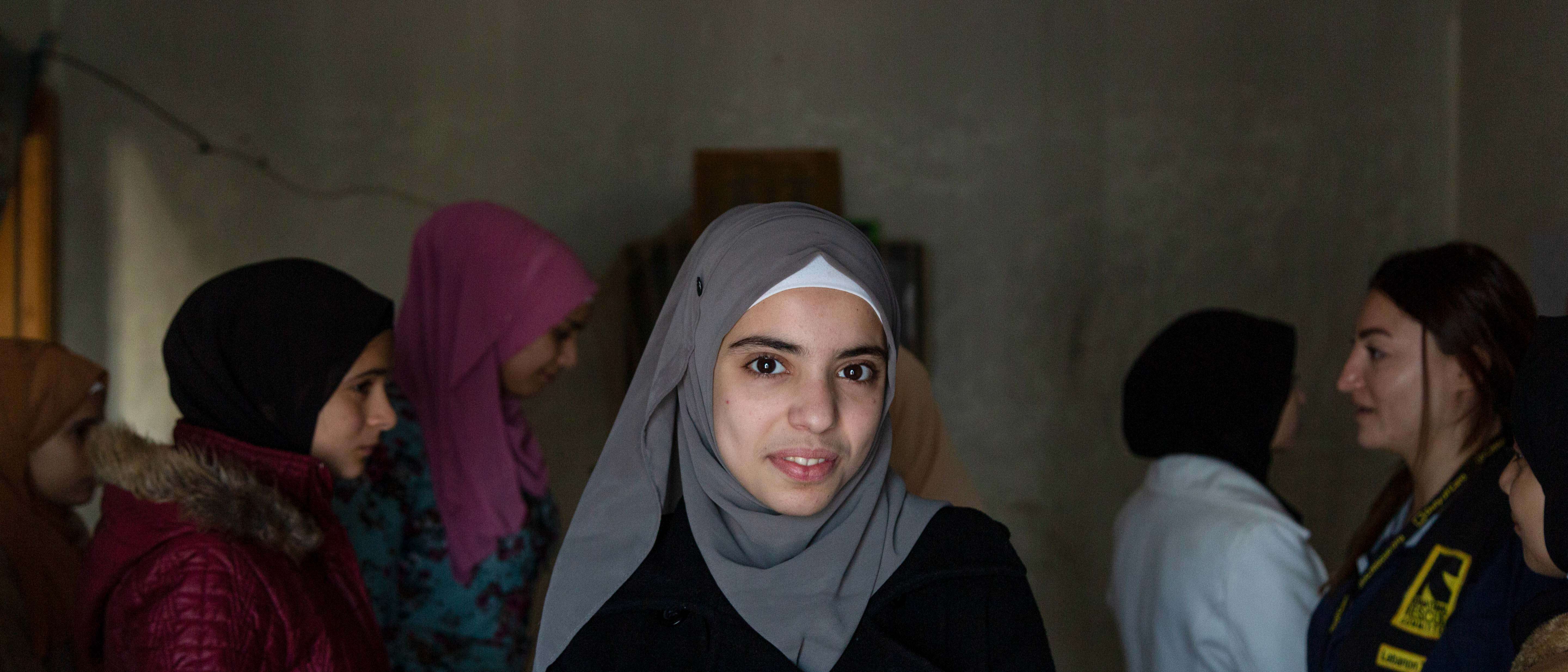 A young woman smiles at the camera in Lebanon. Behind her, other young women speak to an IRC staff member.