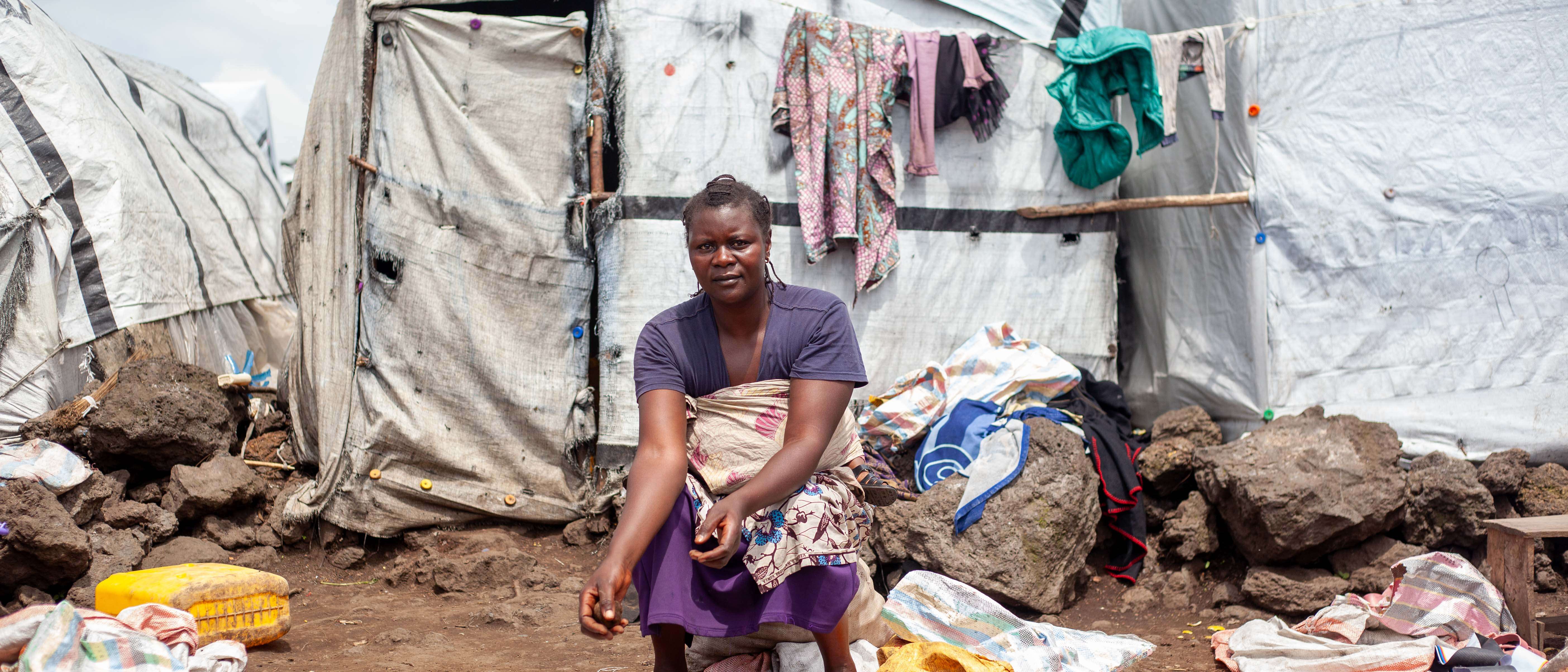 A woman sits outside, in front of a white makeshift shelter. A large pile of potatoes lie in front of her.